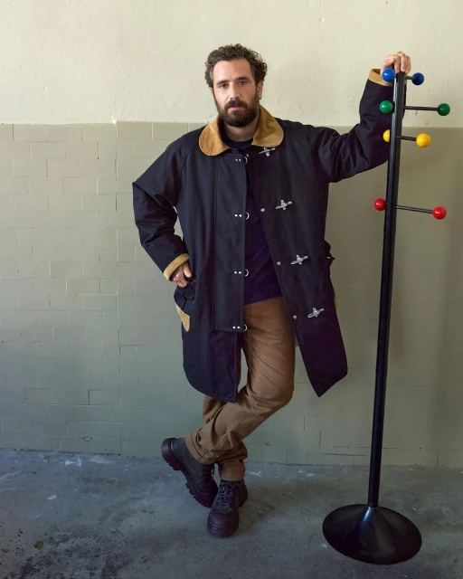 Adult man with a beard wearing a black toggle coat leans against a colorful coat rack inside a room with a concrete floor and wall.