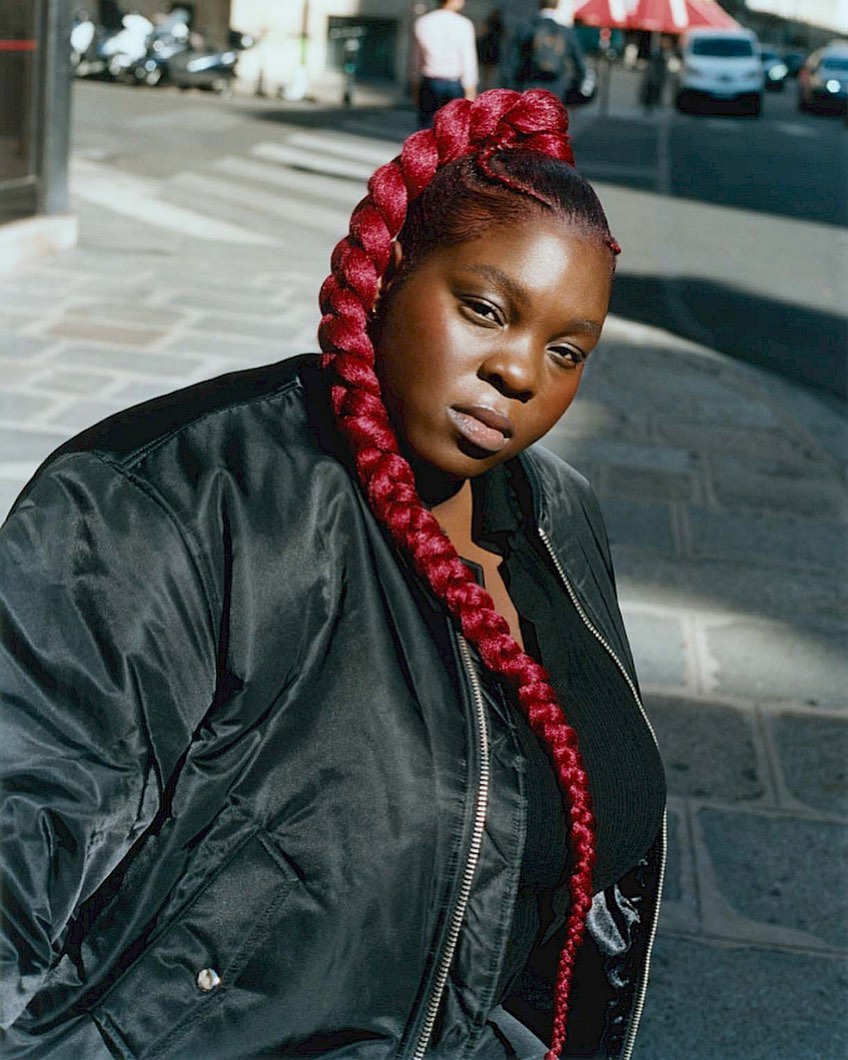 Woman with a long red braided ponytail in an outdoor setting on a sunny day, wearing a black jacket and top. Urban environment.