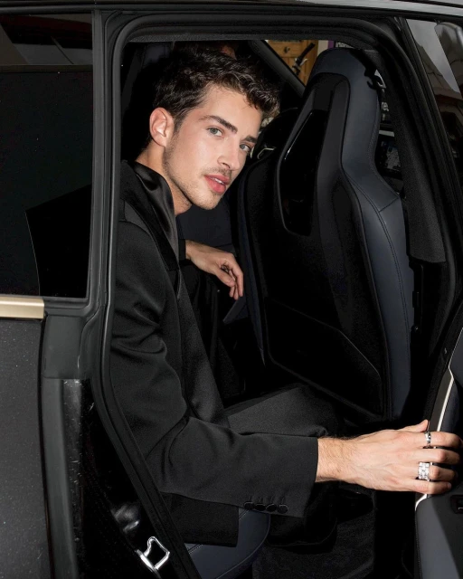 Young man in a black suit poses in the backseat of a car, looking directly at the camera with a serious expression, accessorized with rings.