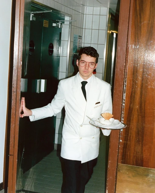 Server stands in a doorway holding a tray with a dessert. He's wearing a white jacket and black tie in an indoor setting.