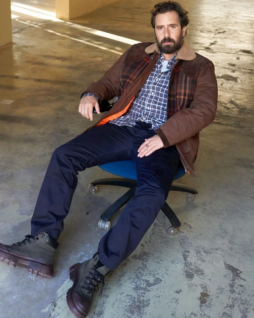 Adult male seated in a chair wearing a brown plaid jacket, blue shirt, and dark pants in a brightly lit indoor setting.