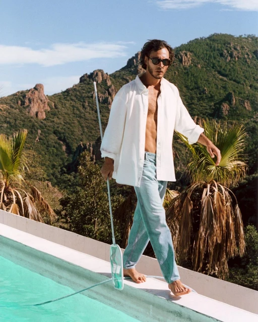 Man balances on pool edge while holding a pool cleaning net, wearing a white shirt and jeans, set against a mountain backdrop under a sunny sky.