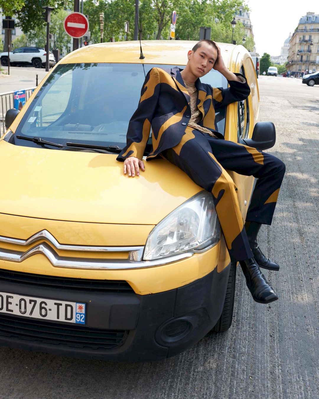 Adult male fashion model in patterned suit poses on yellow Citroen van in Parisian street under blue sky and green trees on a sunny day.