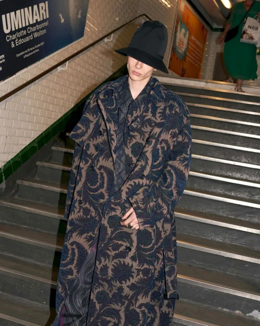 A person wearing a long patterned coat and hat stands on stairs in a subway setting with another person walking behind them.