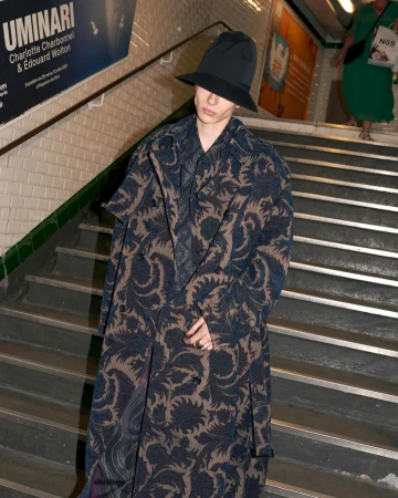 A person wearing a long patterned coat and hat stands on stairs in a subway setting with another person walking behind them.