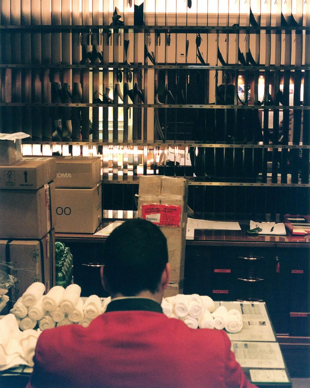 Back view of person in red jacket at service counter with mail slots, stacks of rolled towels, boxes, and papers, indoors.