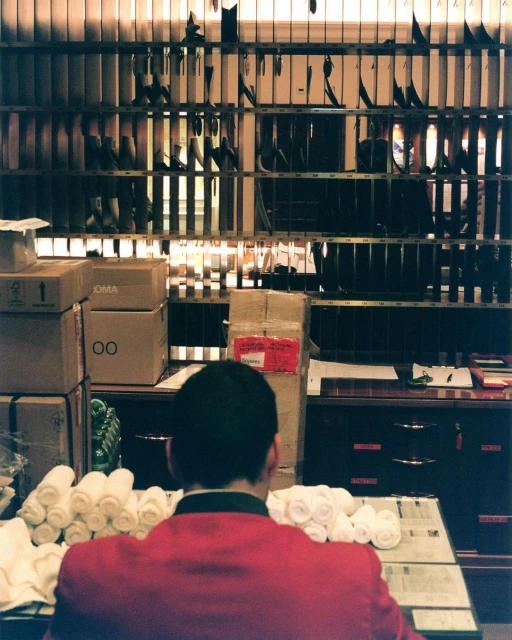 Back view of person in red jacket at service counter with mail slots, stacks of rolled towels, boxes, and papers, indoors.