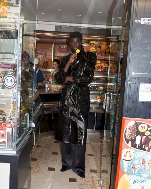 A dark-skinned model in a shiny black coat holds a yellow flower lollipop in front of a bakery storefront with baked goods on display.