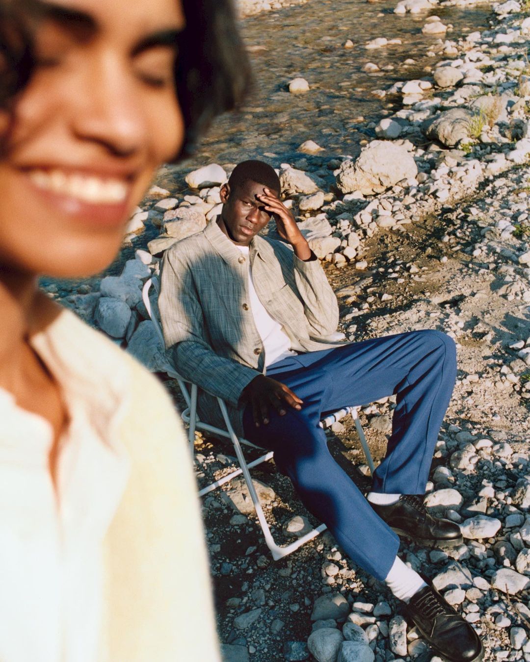 Two people outdoors, one seated in a folding chair with blue pants and the other smiling with a yellow sweater, near a rocky river.