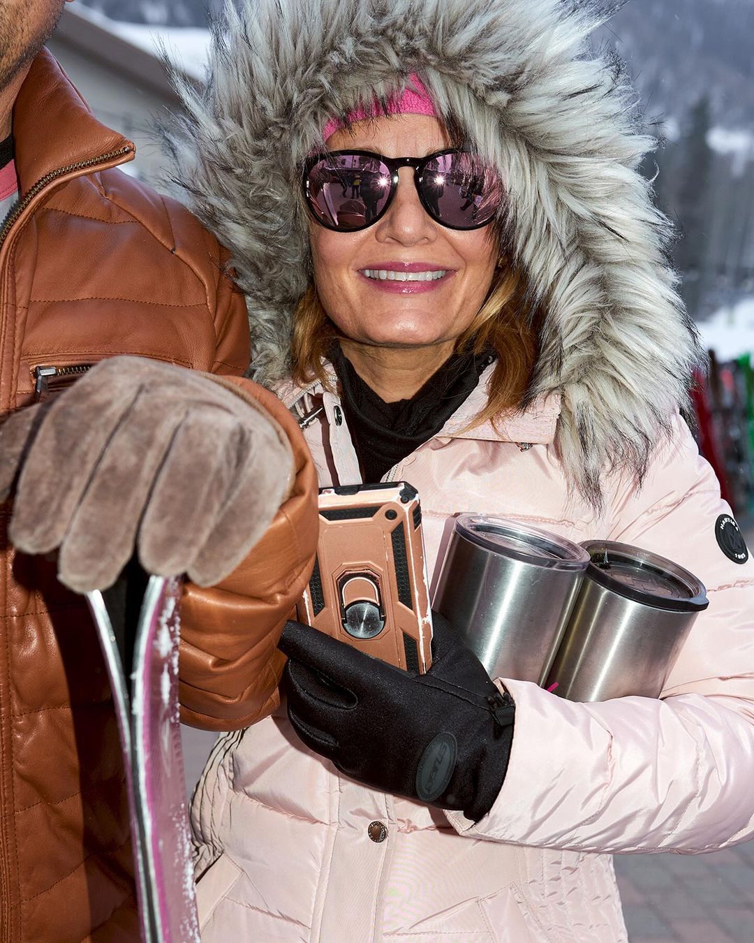 Woman in a fur-trimmed hood and sunglasses, holding stainless steel tumblers, stands next to a man with skis during daytime.