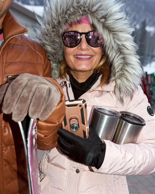 Woman in a fur-trimmed hood and sunglasses, holding stainless steel tumblers, stands next to a man with skis during daytime.