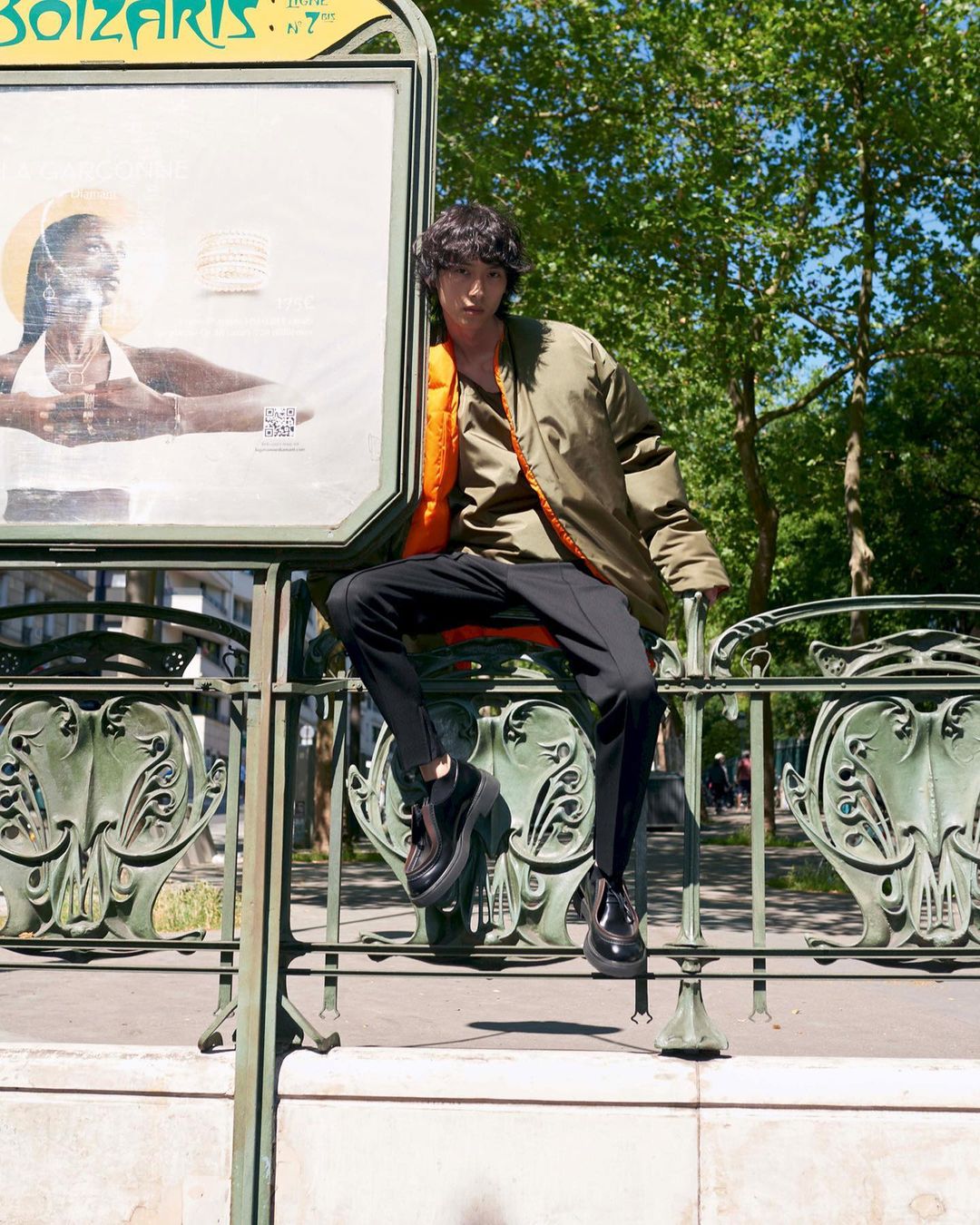 Young Asian man in olive bomber jacket and black pants seated on wrought iron railing of a Paris Metro station in bright sunlight.