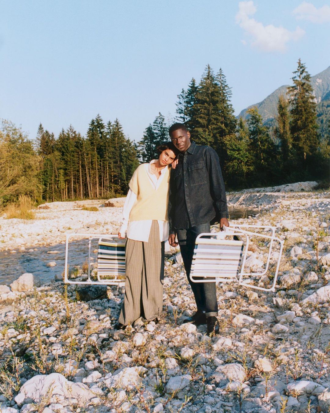 A young couple stands in a rocky riverbed holding beach chairs, with mountains and evergreens in the background under a bright sky.