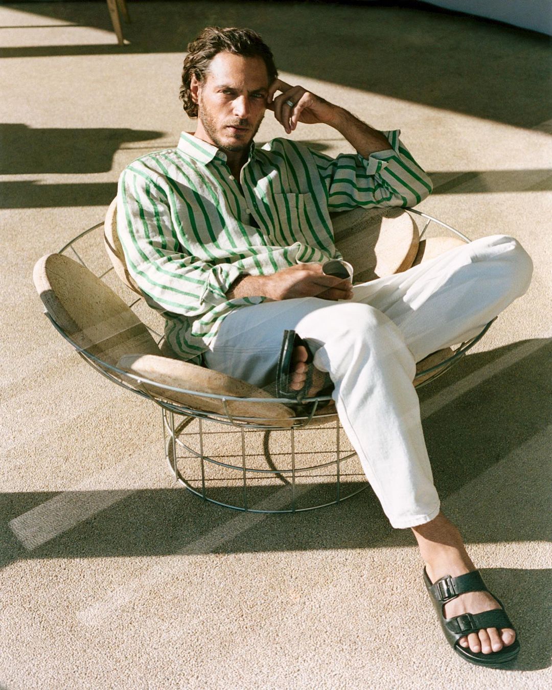 Adult male with striped shirt sits in wire chair with sandals in indoor setting with natural light and neutral color palette.