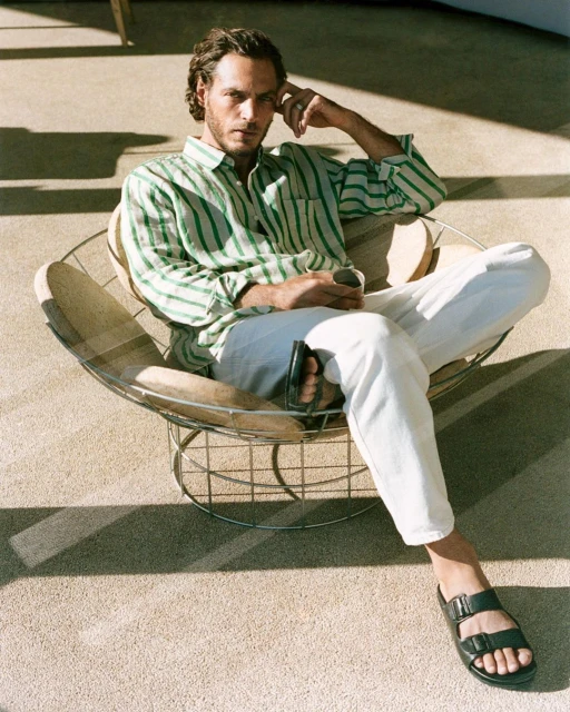 Adult male with striped shirt sits in wire chair with sandals in indoor setting with natural light and neutral color palette.