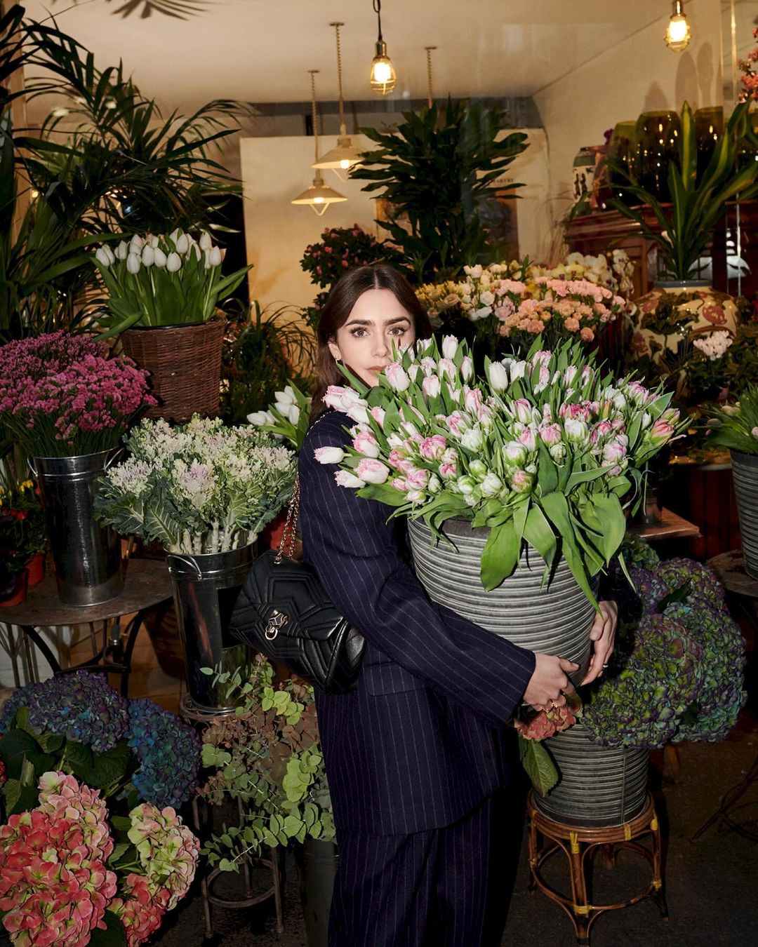 Woman in a pinstripe suit carrying a bucket full of white and pink tulips surrounded by flower displays at a flower shop.