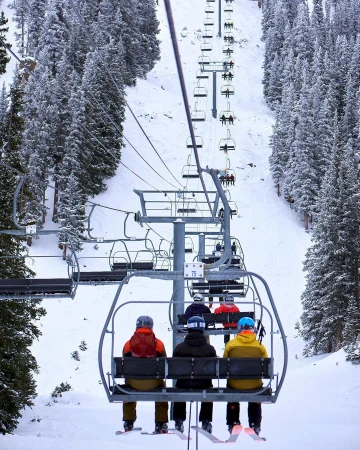 Four skiers ride a chairlift up a snowy mountain. Tall snow-covered evergreen trees and a white ski slope are in the background.