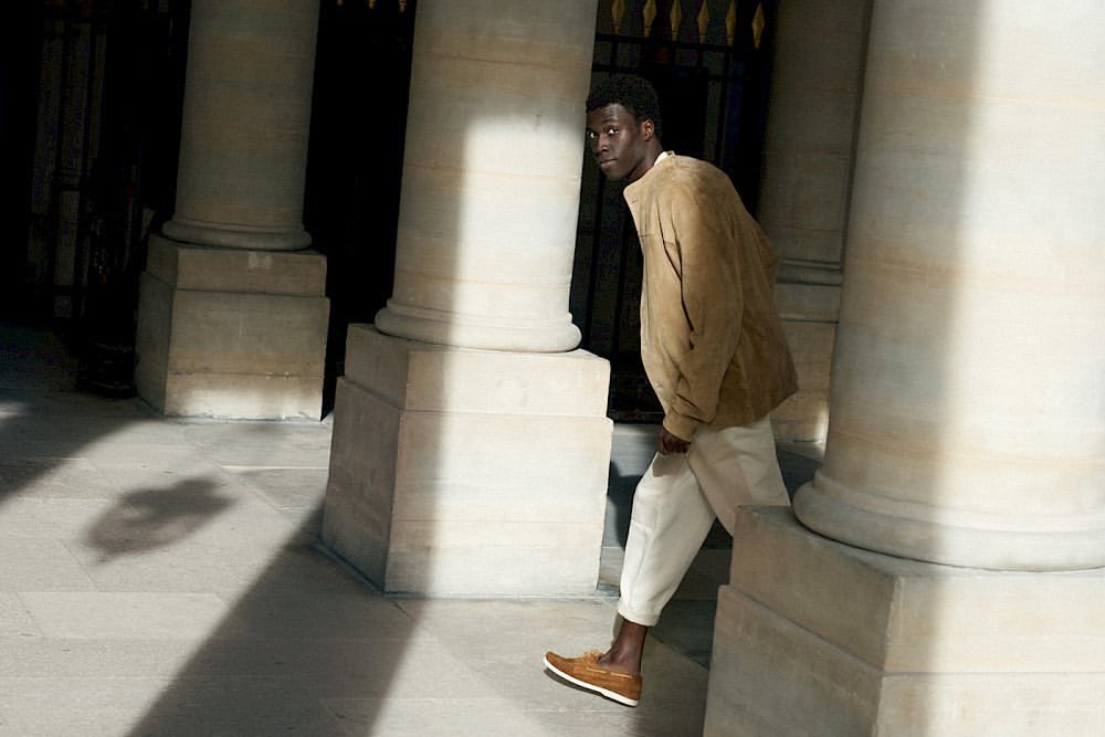Dark-skinned man in a tan jacket and light pants stands between stone columns, illuminated by sunlight on the stone ground.
