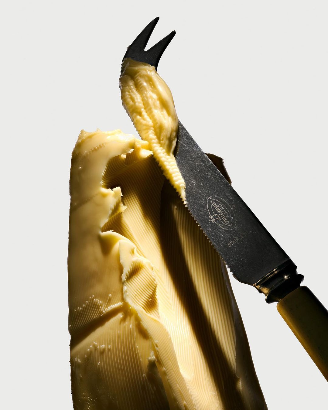 Butter being cut with a butter knife featuring a forked tip, against a white background. The focus is on the texture and form.