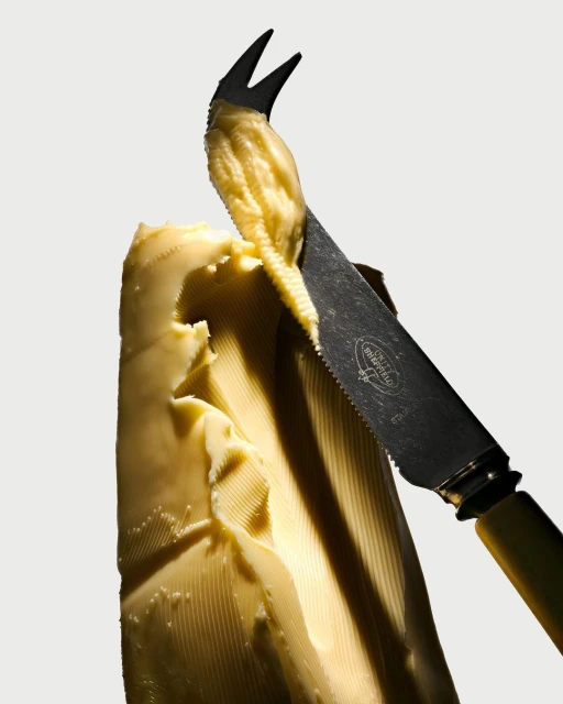 Butter being cut with a butter knife featuring a forked tip, against a white background. The focus is on the texture and form.