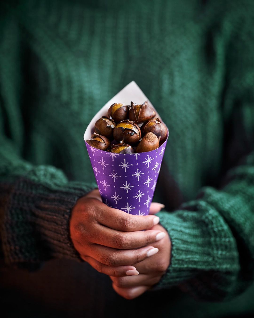 Hands holding a cone with roasted chestnuts. The cone has a purple background and a pattern with white stars. The hands are dark-skinned.
