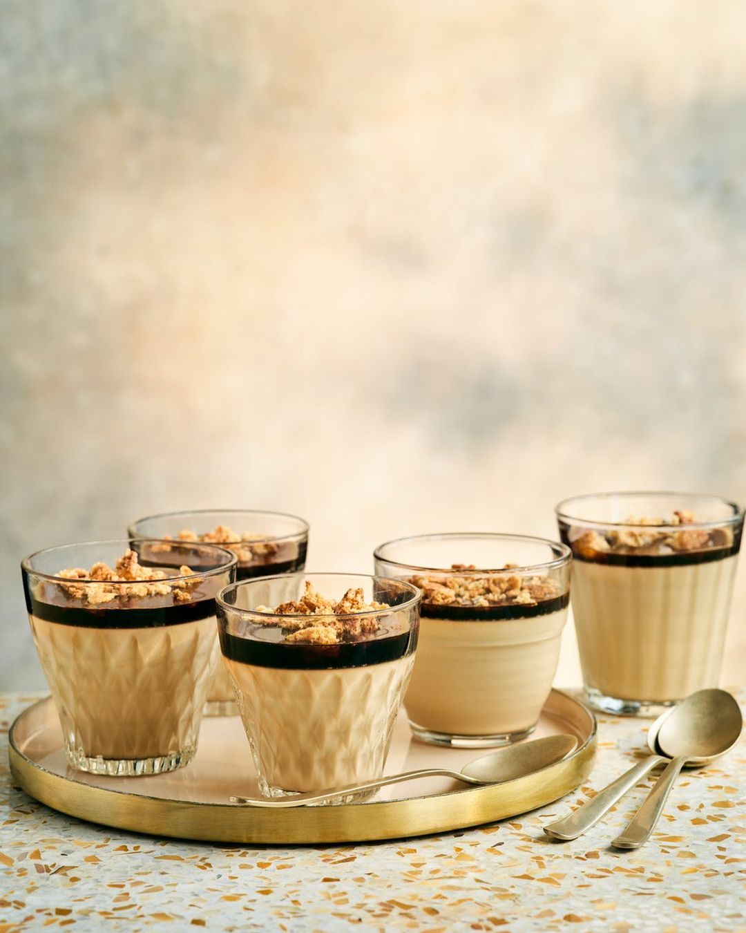 Six dessert cups filled with layered custard, chocolate, and granola topping are arranged on a gold tray with two spoons against a mottled backdrop.