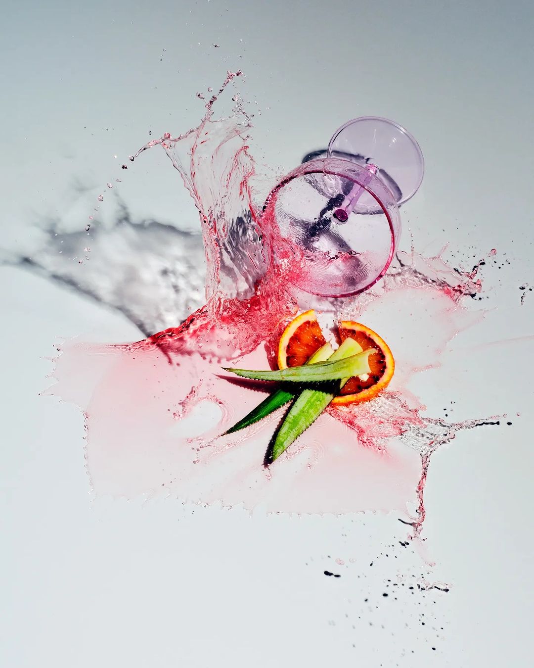 Artistic still life of a cocktail glass with red liquid splashing onto a sliced orange and green leaves against a white background.