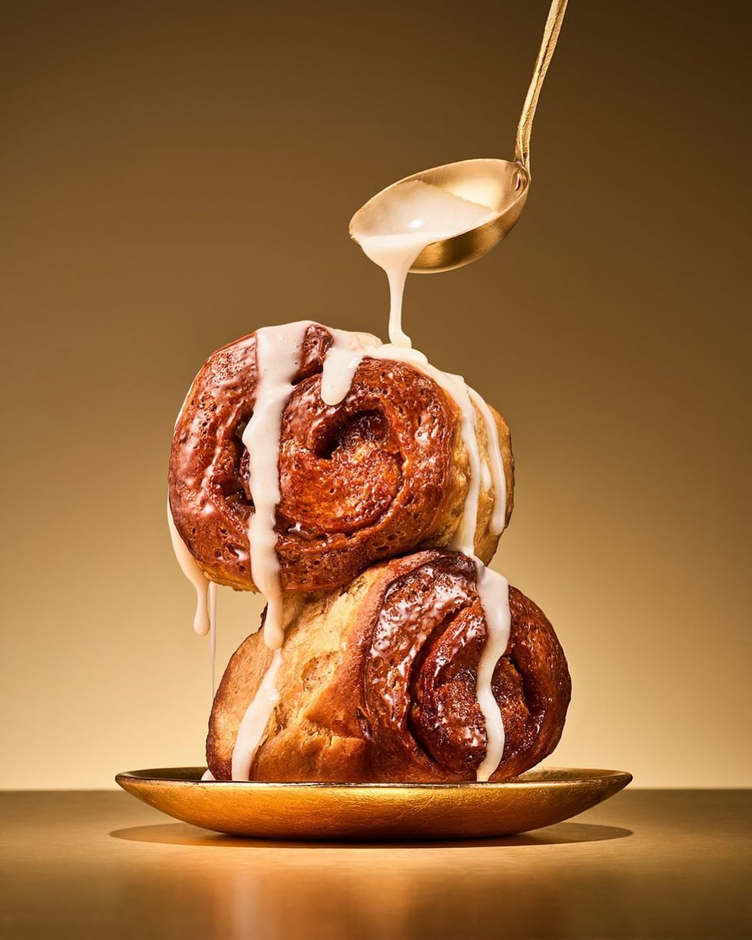 Two golden brown cinnamon rolls are stacked with white glaze dripping down from a golden spoon against a gold background.