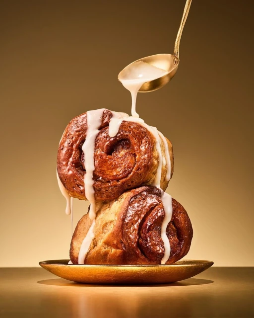 Two golden brown cinnamon rolls are stacked with white glaze dripping down from a golden spoon against a gold background.