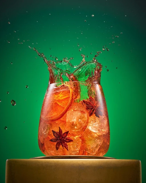Cocktail in a glass with orange slices, ice, and star anise is splashed on a golden base against a green background. 