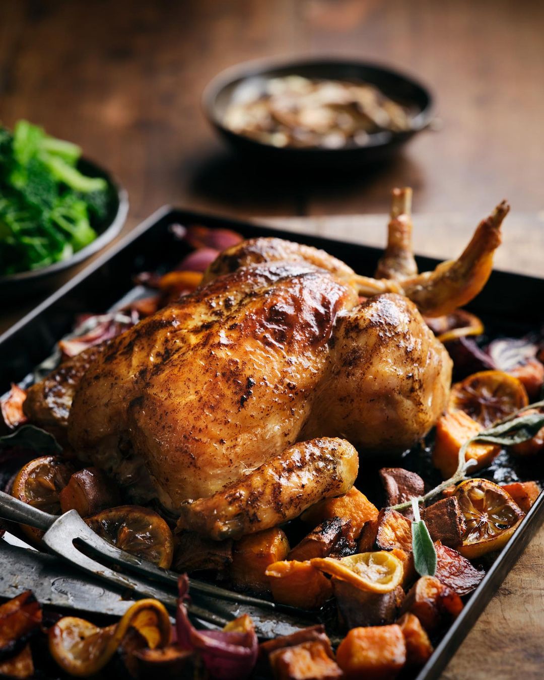 Roasted chicken on a black baking tray, surrounded by vegetables and lemon slices, beside bowls of salad on a wooden surface.