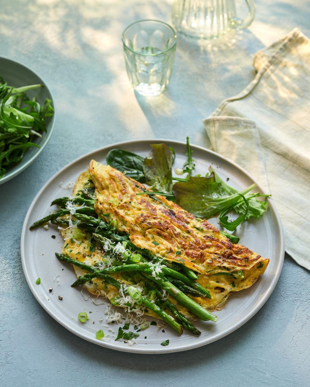 Overhead shot of a breakfast omelet with asparagus spears, Parmesan cheese, and a side salad on a rustic blue table with sunlight.