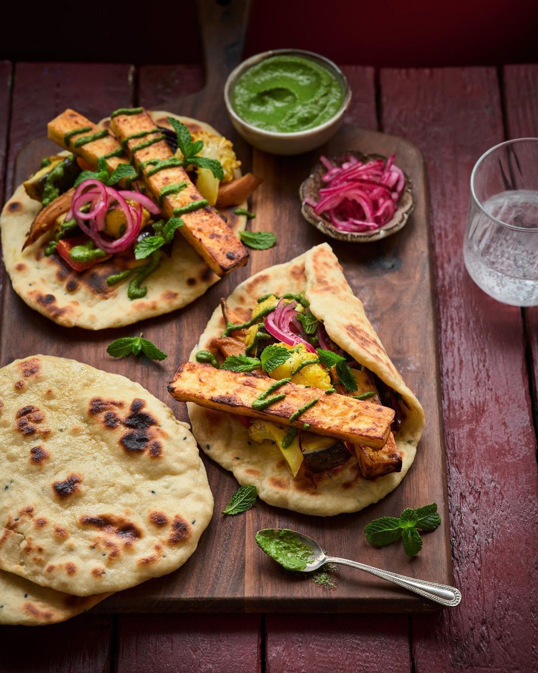 Overhead shot of naan bread topped with paneer, vegetables, and mint, on a wooden board with dips and a glass of water.