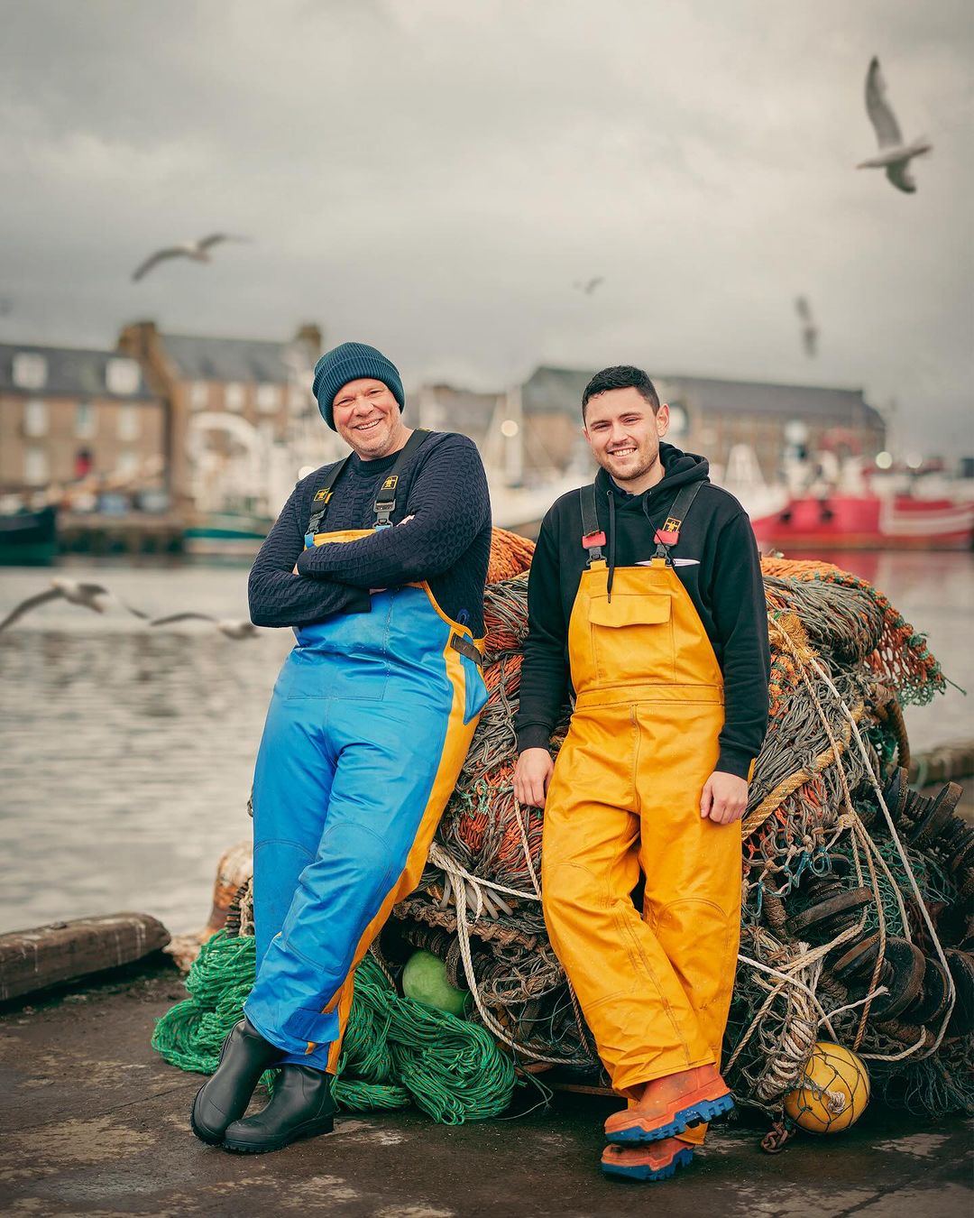 Two fishermen in yellow and blue overalls stand smiling near a harbor on an overcast day. Fishing nets and boats in background.