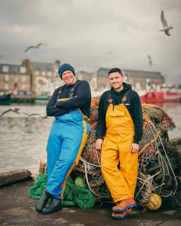 Two fishermen in yellow and blue overalls stand smiling near a harbor on an overcast day. Fishing nets and boats in background.