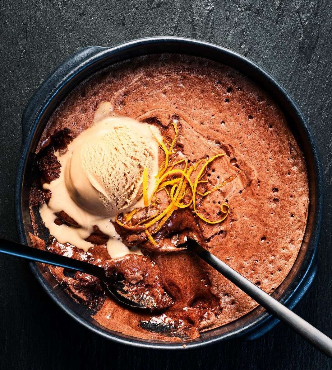 Overhead shot of chocolate lava cake in a dark round baking dish, topped with vanilla ice cream and orange zest, served with spoons.