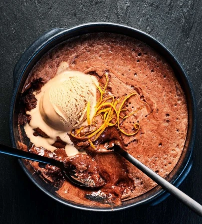 Overhead shot of chocolate lava cake in a dark round baking dish, topped with vanilla ice cream and orange zest, served with spoons.