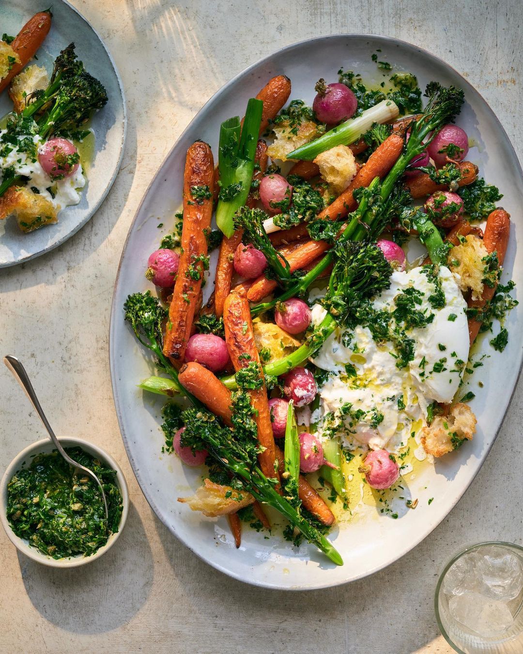 Overhead shot of two plates with vibrant salad of roasted carrots, radishes, broccolini, herbs, and poached egg; a small bowl of pesto.