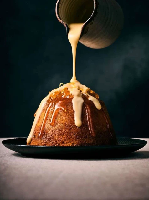 Close-up of a domed cake on a black plate with caramel sauce being poured, set against a moody dark background.