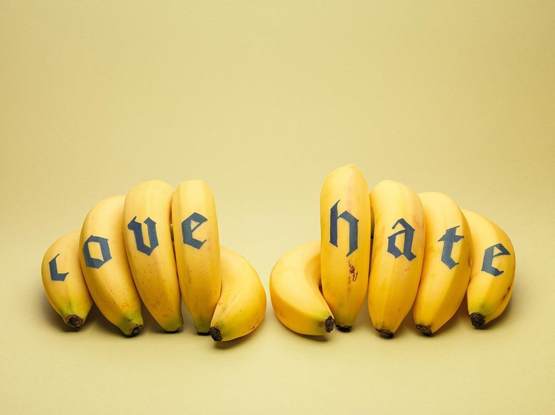 Arrangement of bananas with 'love' and 'hate' written on them, set against a pale yellow backdrop in a studio setting.