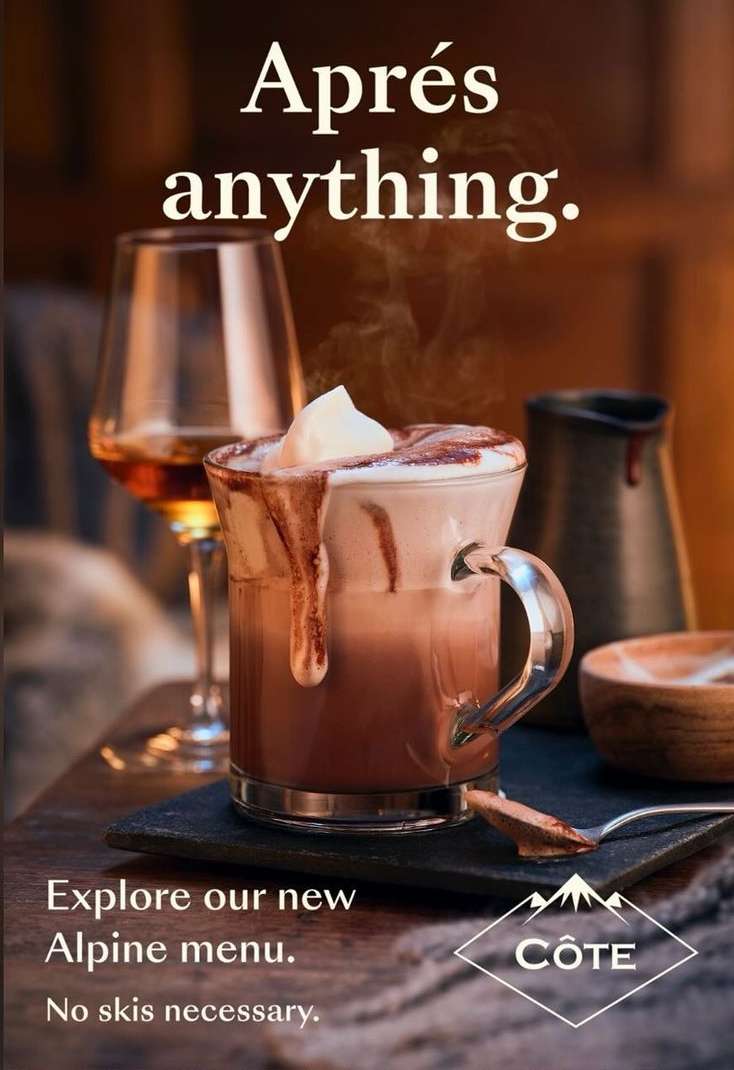 Hot chocolate with whipped cream next to glass of liquor on slate surface, warm lighting, menu visible in a warm indoor environment.