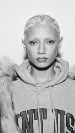 Black and white portrait of a young adult woman with blonde short hair wearing a hoodie with fur trim on a white background.