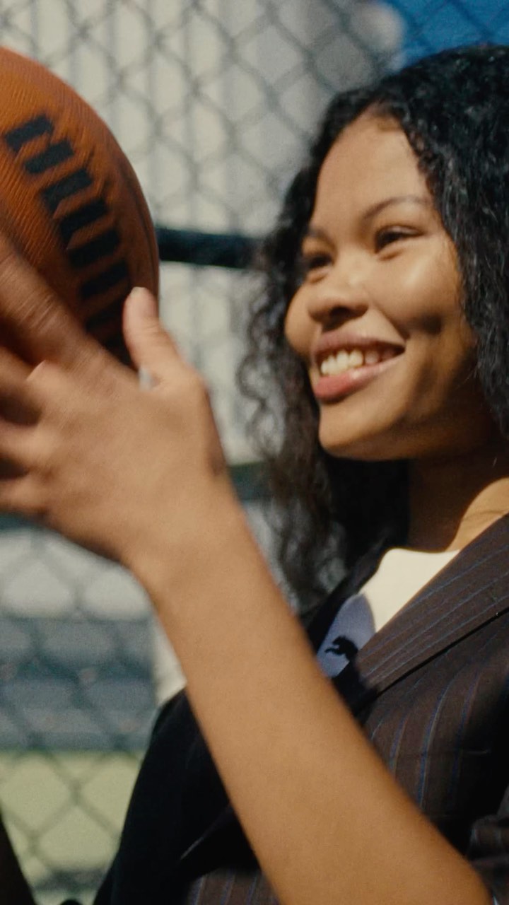 Smiling young woman in a pinstripe suit holding an American football outdoors, with a chain-link fence and blue sky in the background.