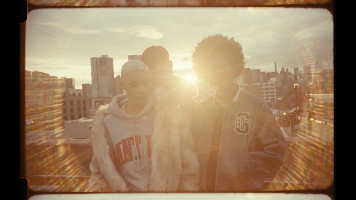 Three people stand on a rooftop overlooking a cityscape bathed in golden light from the setting sun; urban fashion style.
