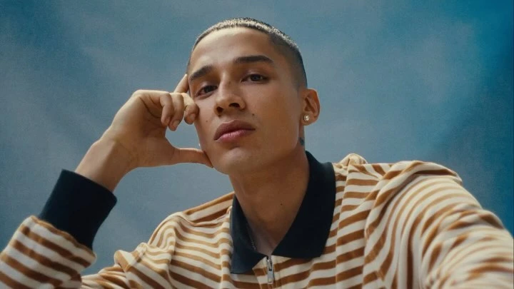 Portrait of a young adult male with short hair, wearing a striped shirt, resting his head on his hand against a blurred blue background.