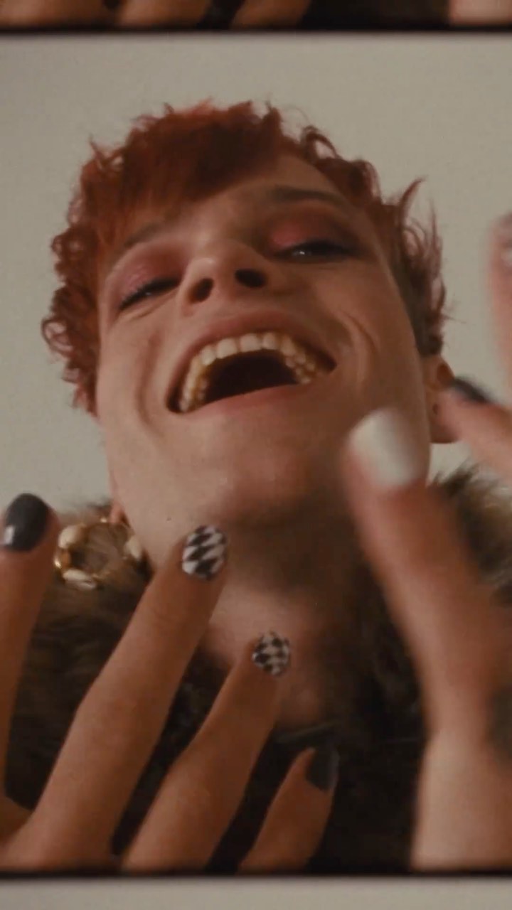 Eye-level close-up of a smiling red-haired woman with open mouth and decorated nails, wearing a fur coat and gold earrings against an indoor background.