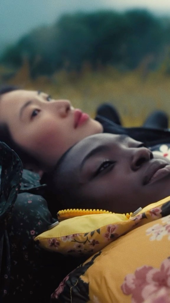 Two women lie side-by-side on a yellow floral patterned pillow outdoors with a blurred background of green foliage and overcast sky.