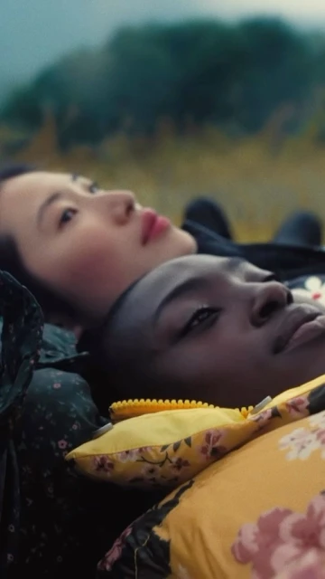 Two women lie side-by-side on a yellow floral patterned pillow outdoors with a blurred background of green foliage and overcast sky.