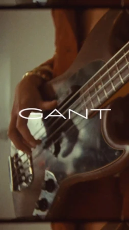 Close-up of a musician playing an electric bass guitar with the brand name GANT overlaid, shot with a shallow depth of field.