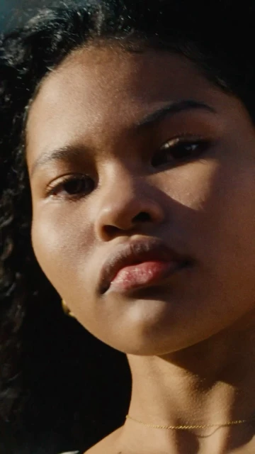 Close-up portrait of a woman with curly black hair, wearing a gold necklace, illuminated by sunlight, creating shadows on her face.